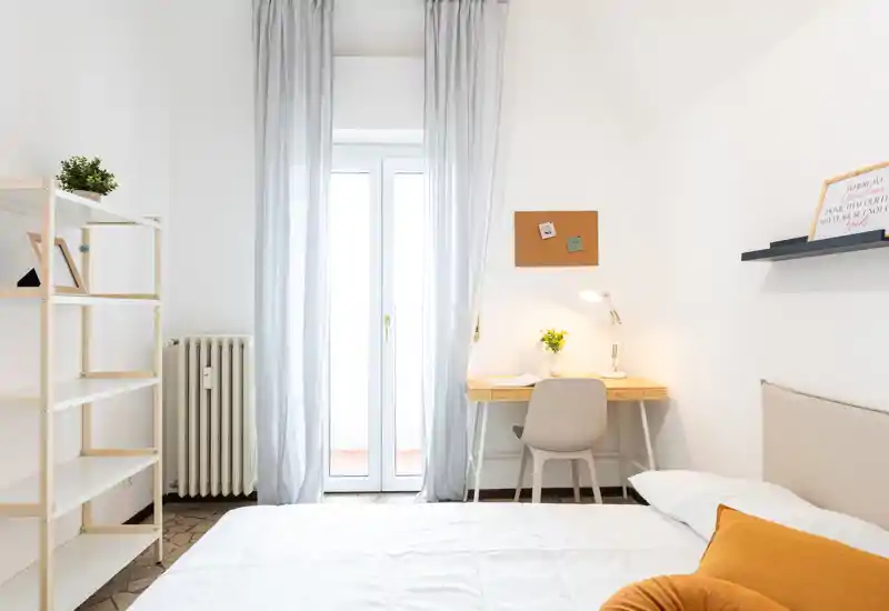 Bright, minimal bedroom with a neatly made bed, soft textiles and a small desk by a tall window that fills the room with natural light.