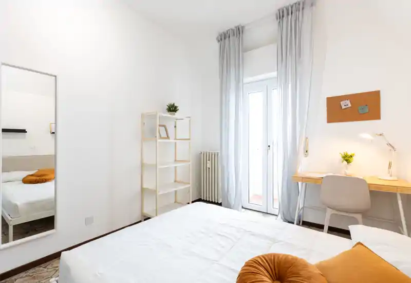 Bright, modern bedroom with a neatly made bed, soft white linens, decorative rust-colored cushions, a shelving unit, and a small work desk by a glass door leading to a balcony.