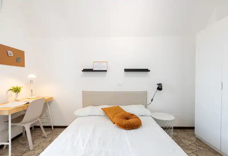 Bright, neatly staged bedroom with a comfortable double bed, warm accent pillow and a small workspace to the side — ideal for a cozy stay.