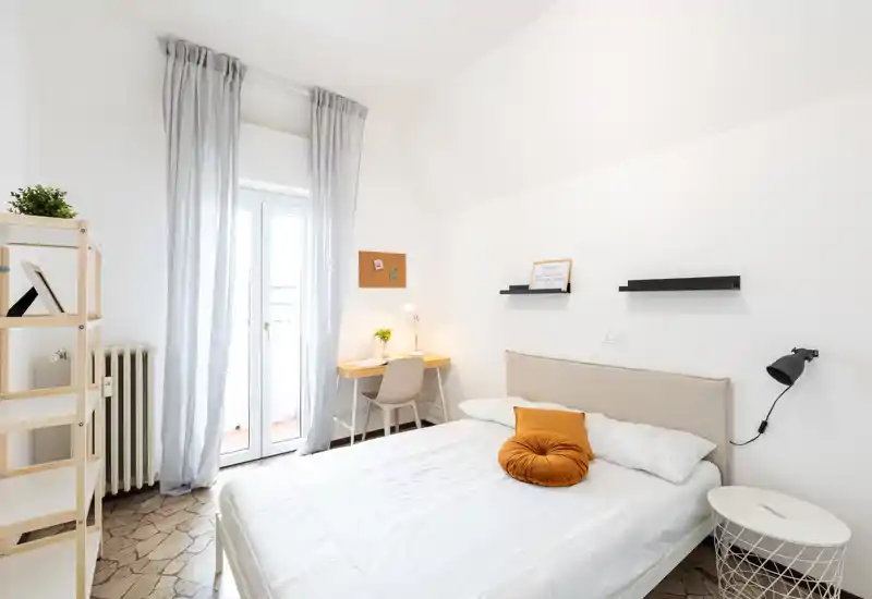 Bright, minimal bedroom with a neatly made double bed, soft headboard and a cozy decorative cushion; a small desk sits beside a tall window/door that opens to a balcony, creating a light and airy atmosphere.
