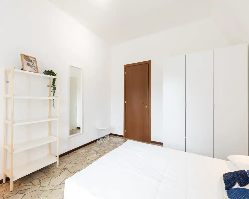 Camera da letto minimalista e luminosa con armadio bianco, mensole chiare e letto ben rifatto in primo piano; toni neutri e luce naturale creano un'atmosfera calma.
