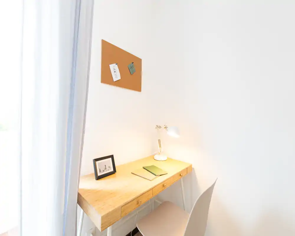 Angolo studio luminoso e minimal con scrivania in legno, lampada da tavolo, pannello in sughero e sedia semplice — perfetto per lavorare in un piccolo spazio abitativo.