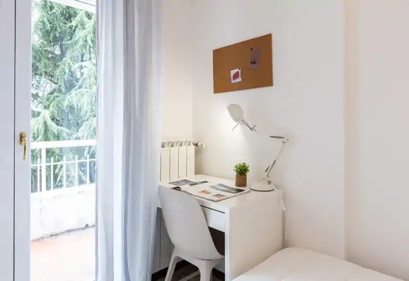 Compact bedroom corner with a small white desk and chair beside a balcony door; soft natural light, a desk lamp and a corkboard create a tidy, minimal workspace next to the bed.