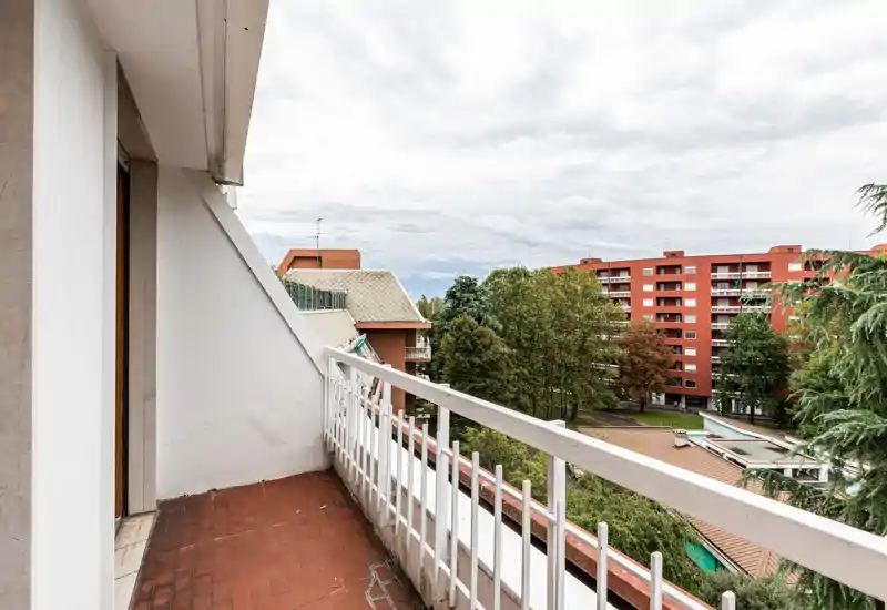 Sunny-feeling balcony with terracotta floor tiles and a white railing, overlooking trees and neighboring apartment blocks — suitable for a small table and chairs.