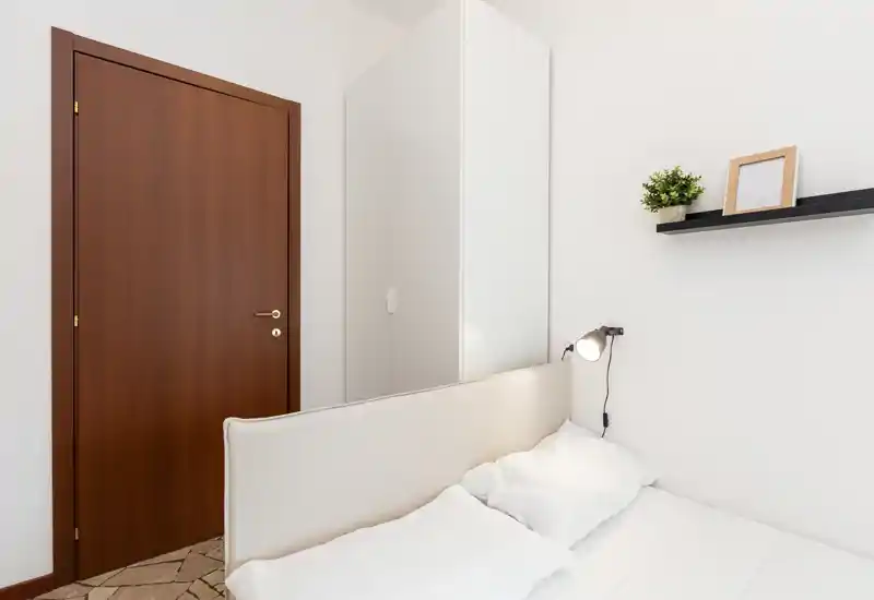 Bright, minimalist bedroom featuring a neatly made double bed with headboard, a wall-mounted reading lamp and a white wardrobe. Neutral tones and simple decor create a calm, modern feel.
