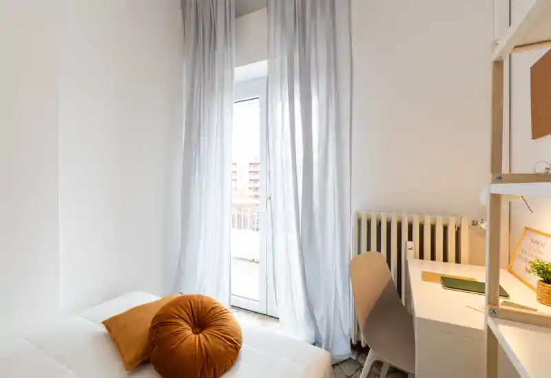 Bright, minimal bedroom with a neatly made bed, warm accent cushions and a desk by the window opening to a small balcony — an airy and functional sleeping corner.