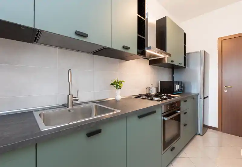 Modern, compact kitchen with a stainless sink, built-in oven and gas hob, matte green cabinets and a tall fridge — bright and well-organized space ideal for short-term rentals.