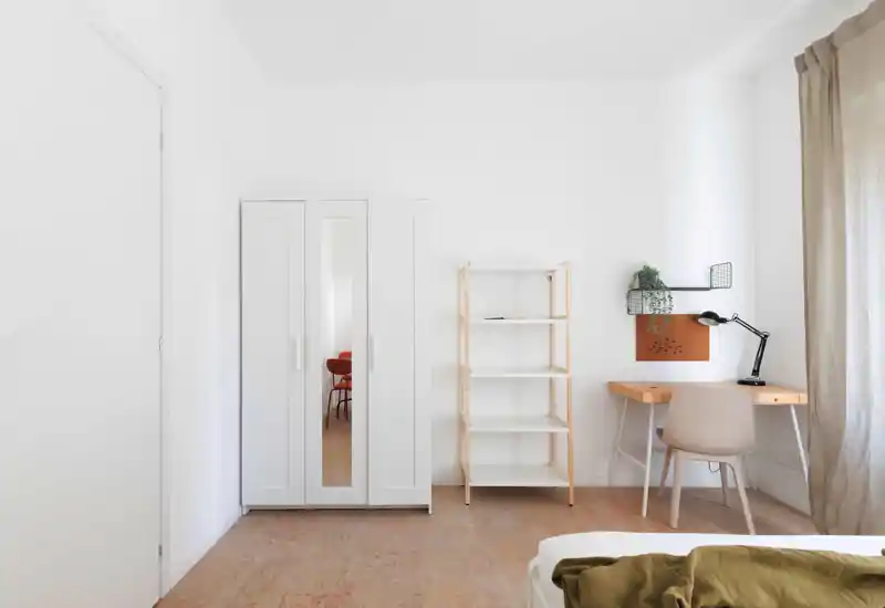 Rincón de dormitorio luminoso y minimalista con escritorio, armario y cama parcialmente visible; paredes blancas y luz natural que aportan una sensación moderna y aireada.