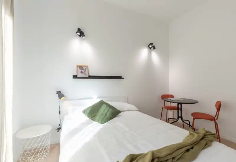 Dormitorio luminoso y minimalista con cama doble bien hecha, ropa de cama blanca, cojín verde como acento y una pequeña mesa redonda con dos sillas — un espacio aireado y acogedor con luz natural.