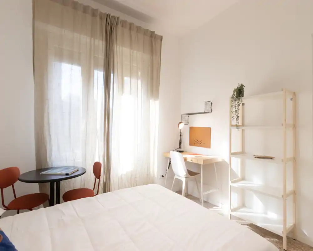 Camera da letto luminosa e minimal con un grande letto in primo piano, una piccola scrivania con lampada sulla destra e una mensola, più un tavolino rotondo con due sedie vicino alla finestra che diffonde luce naturale.