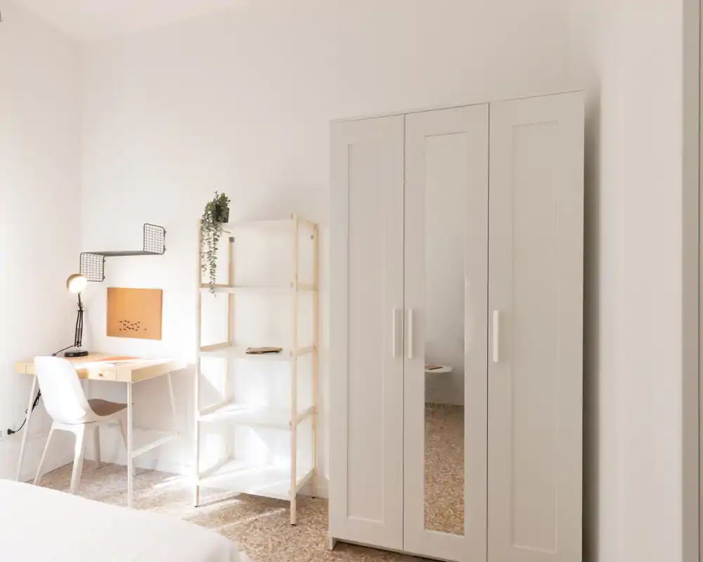 Angolo camera luminoso e minimal con armadio bianco, scaffale e una piccola scrivania in legno — ideale per un soggiorno accogliente e con spazio per lavorare.