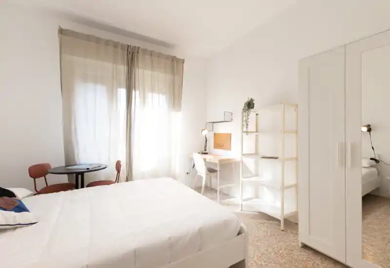Camera da letto luminosa e minimale con ampio letto bianco, scrivania per lo studio e un piccolo angolo conversazione vicino alla finestra con tende leggere. Palette neutra e luce naturale creano un ambiente arioso e accogliente, perfetto per gli annunci.