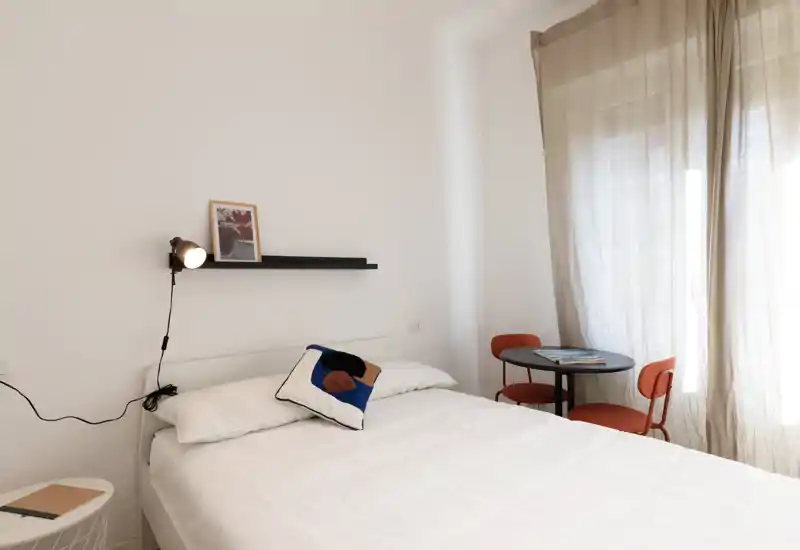 Camera da letto luminosa e minimal con letto matrimoniale con lenzuola bianche, cuscino decorativo, lampada da parete e un piccolo tavolo rotondo con due sedie vicino alla finestra con tenda leggera.