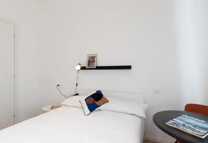 Camera da letto luminosa e minimalista con letto ben rifatto, cuscino decorativo, piccola lampada e comodino; pareti bianche che creano un'atmosfera rilassante.