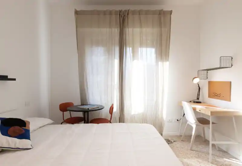 Camera da letto luminosa e minimal con un ampio letto rifatto, un piccolo tavolo rotondo con due sedie vicino alla finestra e una scrivania con lampada — ideale per soggiorni confortevoli.