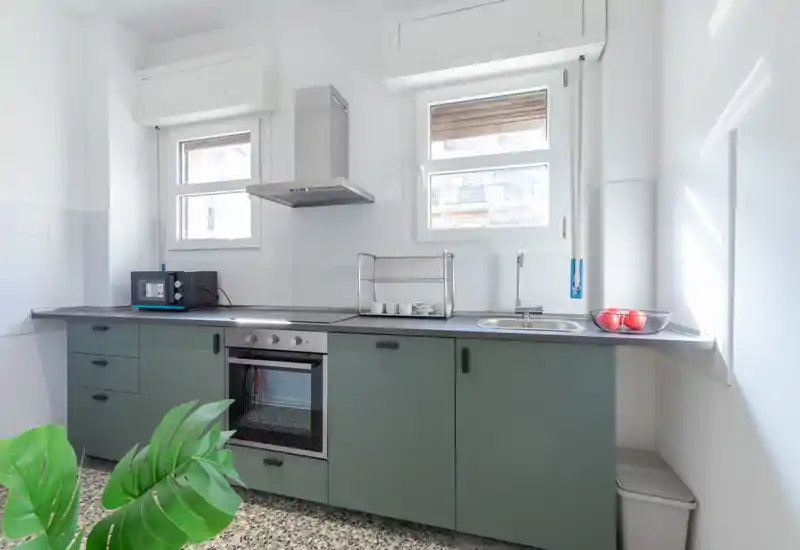 Cocina moderna y luminosa con muebles bajos verdes, horno empotrado, fregadero y dos ventanas que aportan luz natural. Encimeras limpias, un microondas pequeño y una planta decorativa crean un espacio funcional y acogedor.