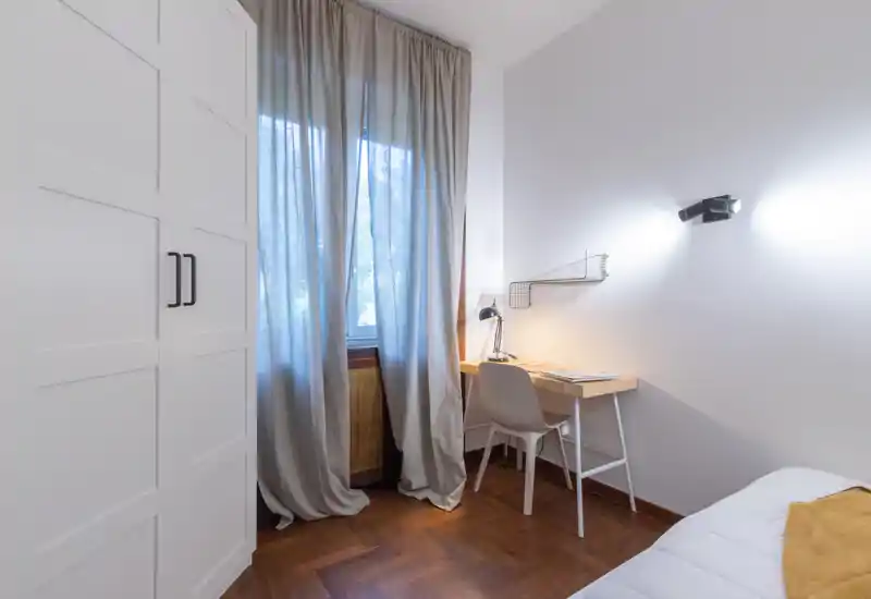 Bright, compact bedroom corner with a small desk, chair and wardrobe; soft curtains and a bedside glimpse of the mattress create a cozy, well-styled space.