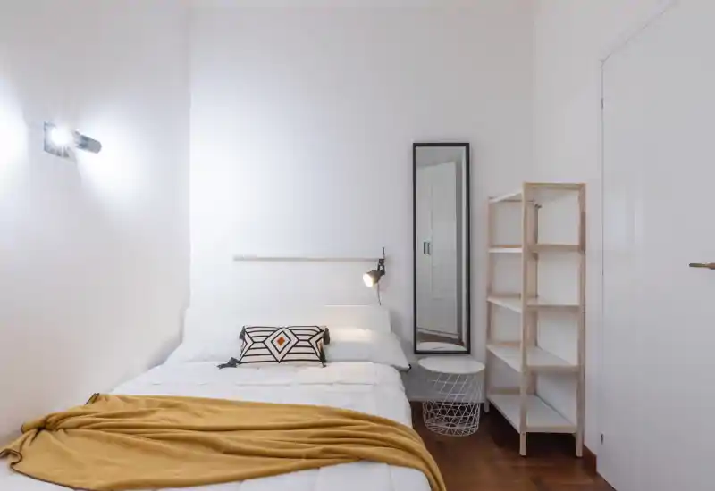 Bright, minimalist bedroom with a neatly made double bed, decorative cushion and a mustard throw; a full-length mirror and open shelving complete the cozy, modern space.