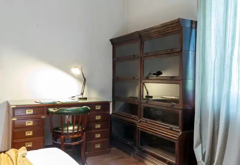 Classic bedroom corner with a wooden desk, green leather chair and a stacked wooden cabinet; soft daylight from a curtained window and a desk lamp create a warm, usable workspace next to the bed.