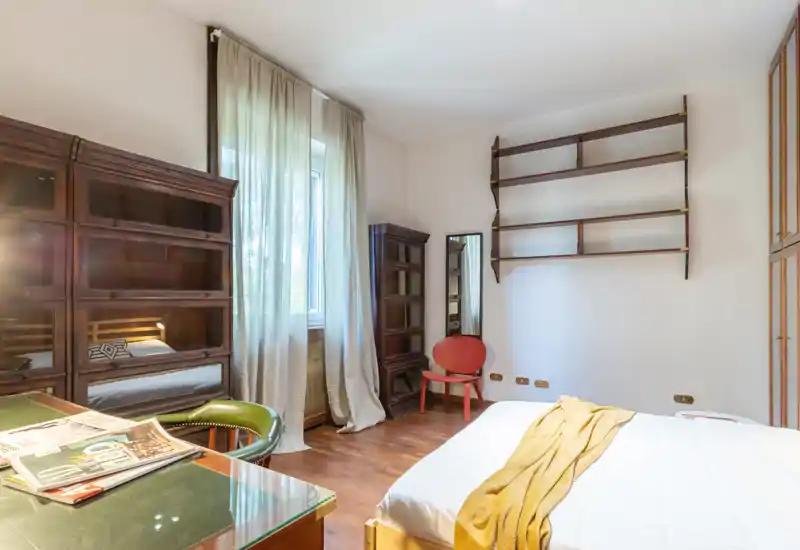 Bright, well-styled bedroom with a visible double bed, wooden flooring and warm vintage shelving; natural light filters through sheer curtains creating a cozy atmosphere.