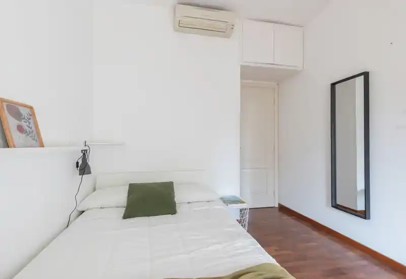 Minimal, bright bedroom with a single bed dressed in white linens, a green cushion, wall-mounted shelf and a full-length mirror. Wood flooring and a neutral palette create a calm, uncluttered space ideal for short-term rentals.