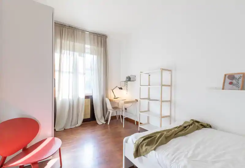 Bright, minimalist bedroom with a single bed, wooden floor and a small desk by the window; soft curtains filter abundant natural light.