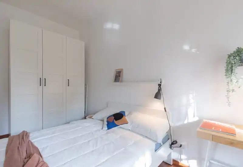 Bright, minimal bedroom with a neatly made double bed, white wardrobe and a small bedside desk. Natural light and simple decor create a calm, airy atmosphere ideal for listing photos.