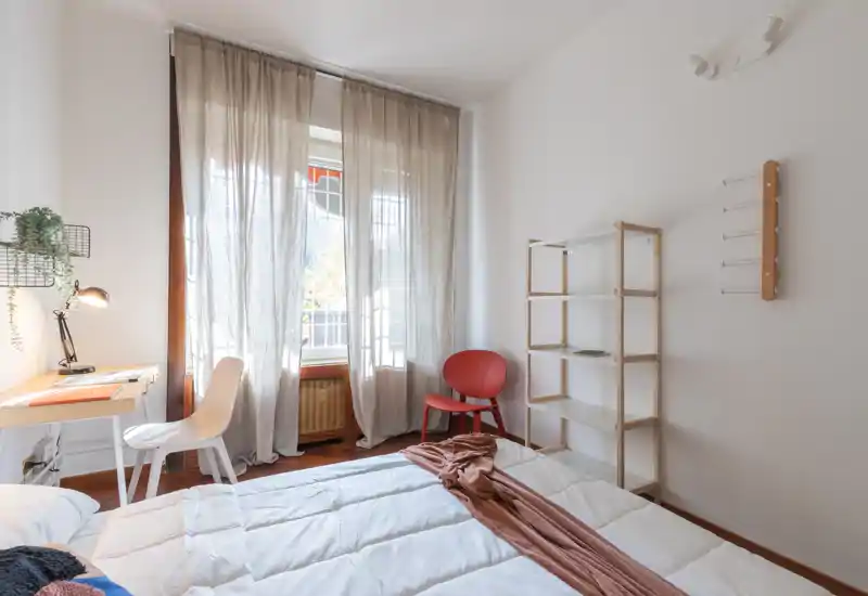 Bright, minimal bedroom with a neatly made bed, soft natural light from a large window, a compact desk and simple shelving — ideal for a cozy single or short-stay rental.