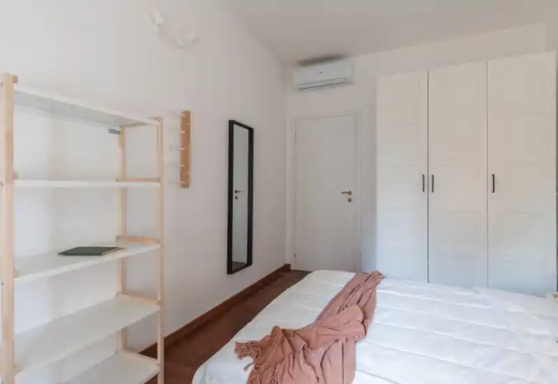 Bright, minimal bedroom with a neatly made bed, wardrobe, wall mirror and shelving — clean lines and soft natural light create an airy, comfortable atmosphere.