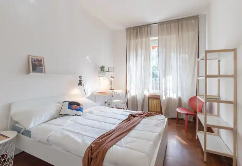 Bright, minimal bedroom with a large double bed, soft linens and a small desk by the window; warm wooden floor and neutral decor create a calm atmosphere.
