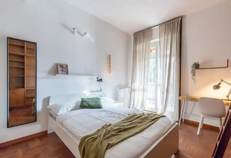 Camera da letto luminosa e ben arredata con letto completo, tessili morbidi e scrivania vicino alla finestra; ampia finestra con tende leggere che dona molta luce naturale.