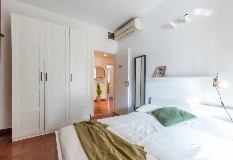 Camera da letto moderna e luminosa con armadio bianco, letto matrimoniale con biancheria bianca e dettagli verdi, lampada a parete e specchio a figura intera — ambiente pulito e arioso.