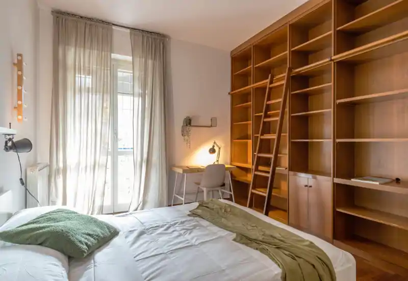Camera da letto luminosa e accogliente con un ampio letto in primo piano, tende leggere che filtrano la luce naturale, una scrivania con lampada e ampie scaffalature in legno con scala.