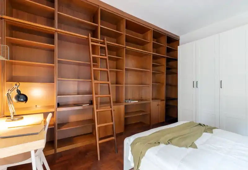 Camera da letto luminosa con una grande libreria in legno integrata e un letto comodo in primo piano. Una scala a pioli aggiunge carattere e un armadio bianco offre spazio extra: linee pulite e toni caldi del legno.