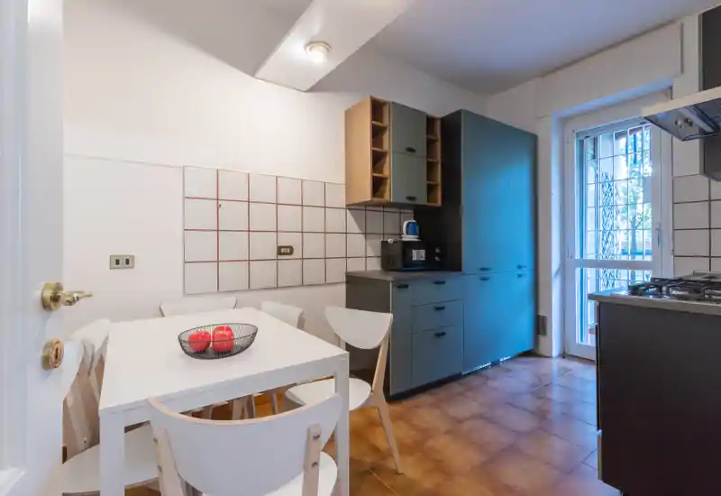 Bright, modern kitchen with a compact dining table, four chairs and built-in cabinets. Natural light enters through a glass door, and the space features a gas hob and cabinetry in muted tones.