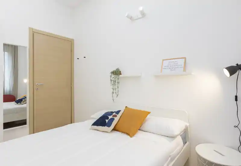 Dormitorio luminoso y minimalista con cama doble cómoda, cojines decorativos y una lámpara de pie. Paredes blancas, estantes sencillos y detalles cálidos que crean un ambiente sereno y acogedor.