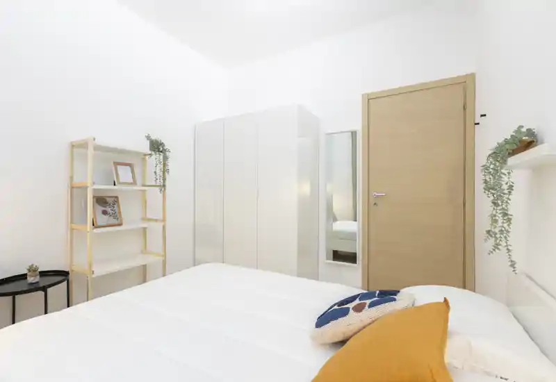 Dormitorio luminoso y minimalista con cama bien hecha, armario blanco y estanterías sencillas; cojines cálidos y plantas añaden encanto.