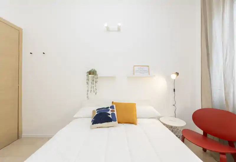 Dormitorio luminoso y minimalista con cama bien hecha, cojines decorativos y una pequeña lámpara de mesilla; estilo limpio y luz natural agradable.