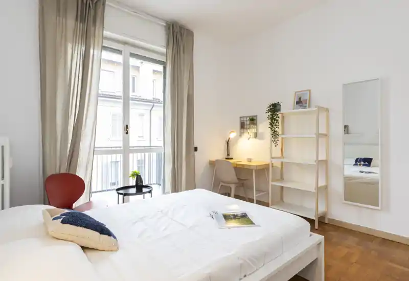 Bright, modern bedroom with a large bed, soft linens and a desk by the window. Neutral tones, a full-length mirror and shelving create a calm, welcoming space ideal for relaxing or working.
