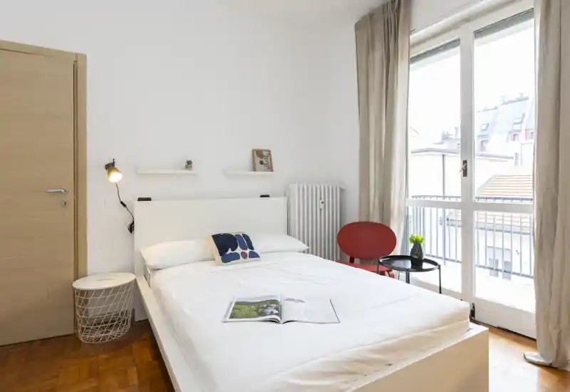 Bright, minimalist bedroom with a white double bed, bedside lamp and balcony doors letting in abundant natural light.