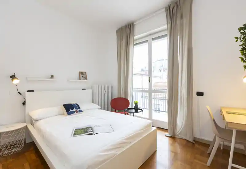 Bright, modern bedroom with a white double bed, bedside lighting, a small seating area and a desk by a large window leading to a balcony.