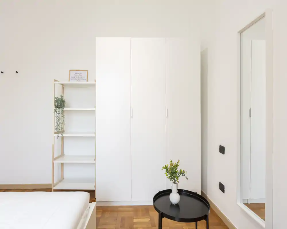 Rincón de dormitorio luminoso y minimalista con armario blanco, borde de la cama visible y una mesa redonda pequeña con un jarrón — limpio, moderno y aireado.