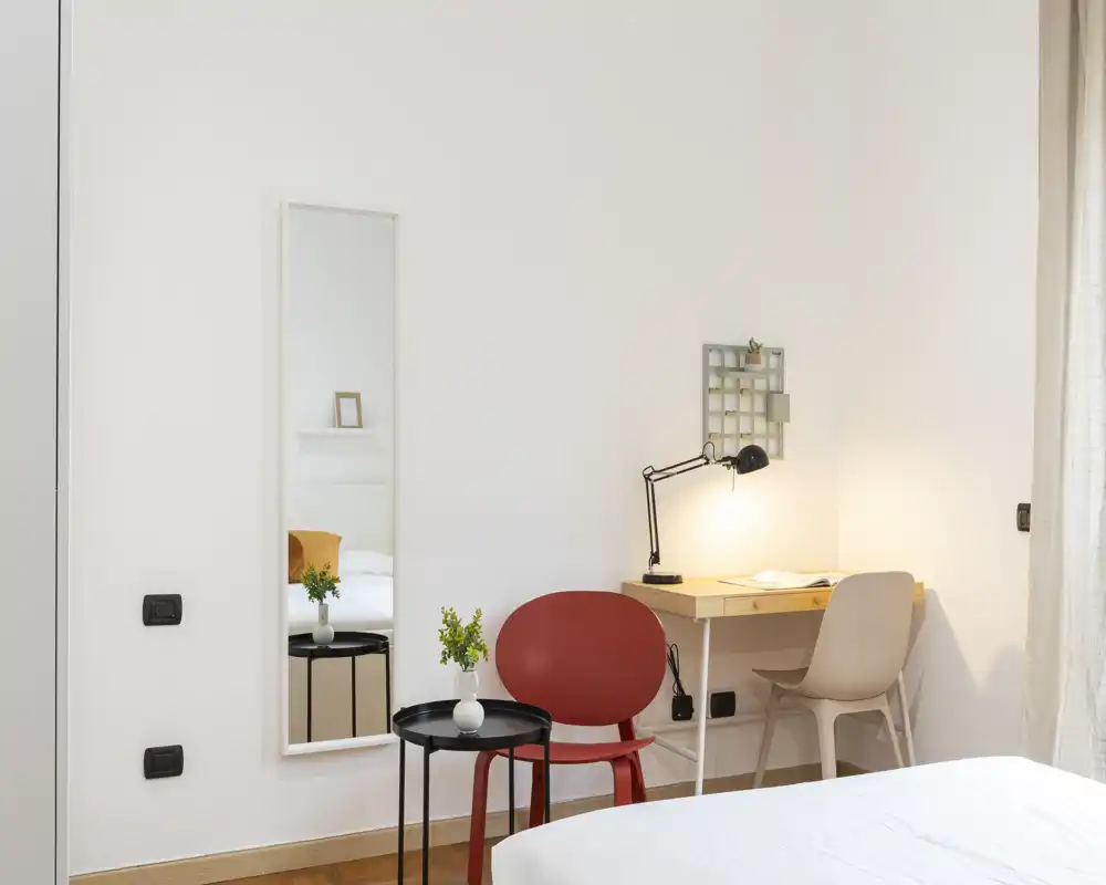 Dormitorio luminoso y minimalista con esquina de la cama visible, escritorio con lámpara y sillas — espacio tranquilo y bien iluminado, perfecto para descansar y trabajar.