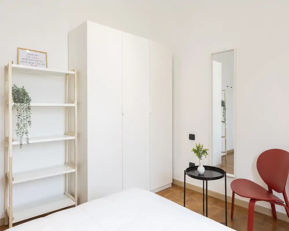 Dormitorio luminoso y minimalista con armario blanco, estantería abierta y una silla roja como acento; la cama se ve parcialmente en primer plano con luz natural y decoración sencilla.