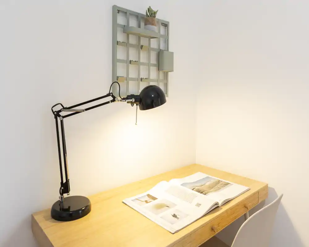 Rincón de escritorio luminoso y minimalista con una lámpara de mesa negra articulada, una revista abierta y una pequeña estantería con una planta — un espacio de trabajo compacto y bien iluminado.
