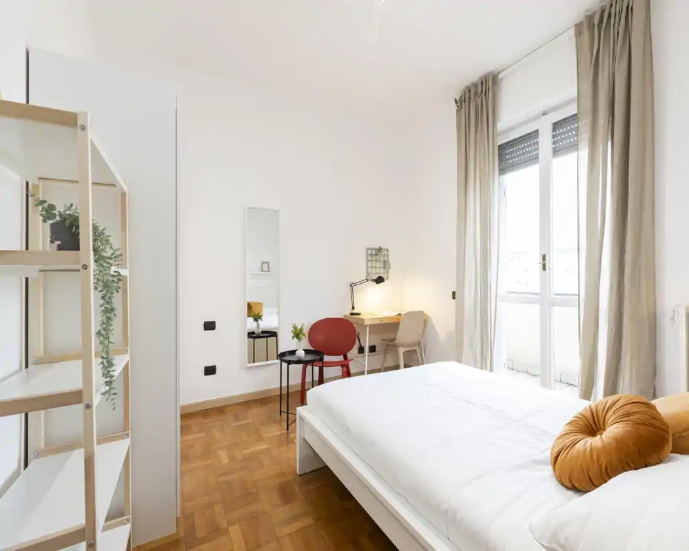 Dormitorio moderno y luminoso con cama blanca, cojín redondo decorativo y mucha luz natural desde la puerta al balcón; incluye un escritorio pequeño y zona de asientos.