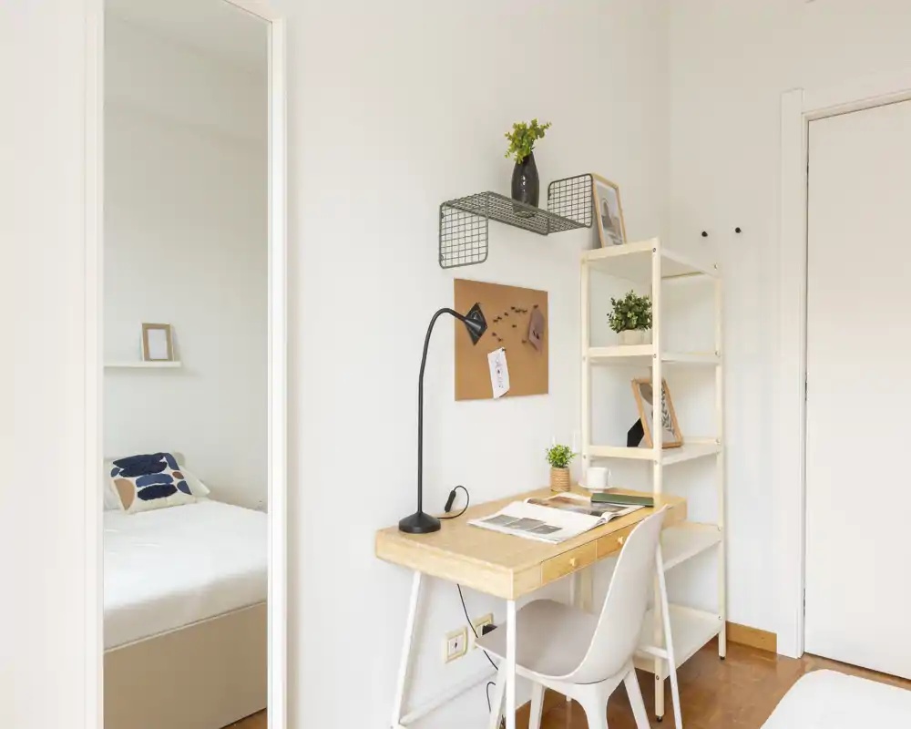 Bright, minimal bedroom corner with a visible bed, a wooden desk and shelving. Natural light and simple décor create a calm, functional workspace next to the sleeping area.