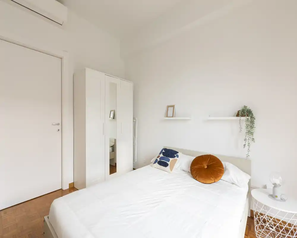Bright, minimal bedroom with a neatly made double bed, decorative pillows and a white wardrobe; calm, modern styling ideal for rental listings.