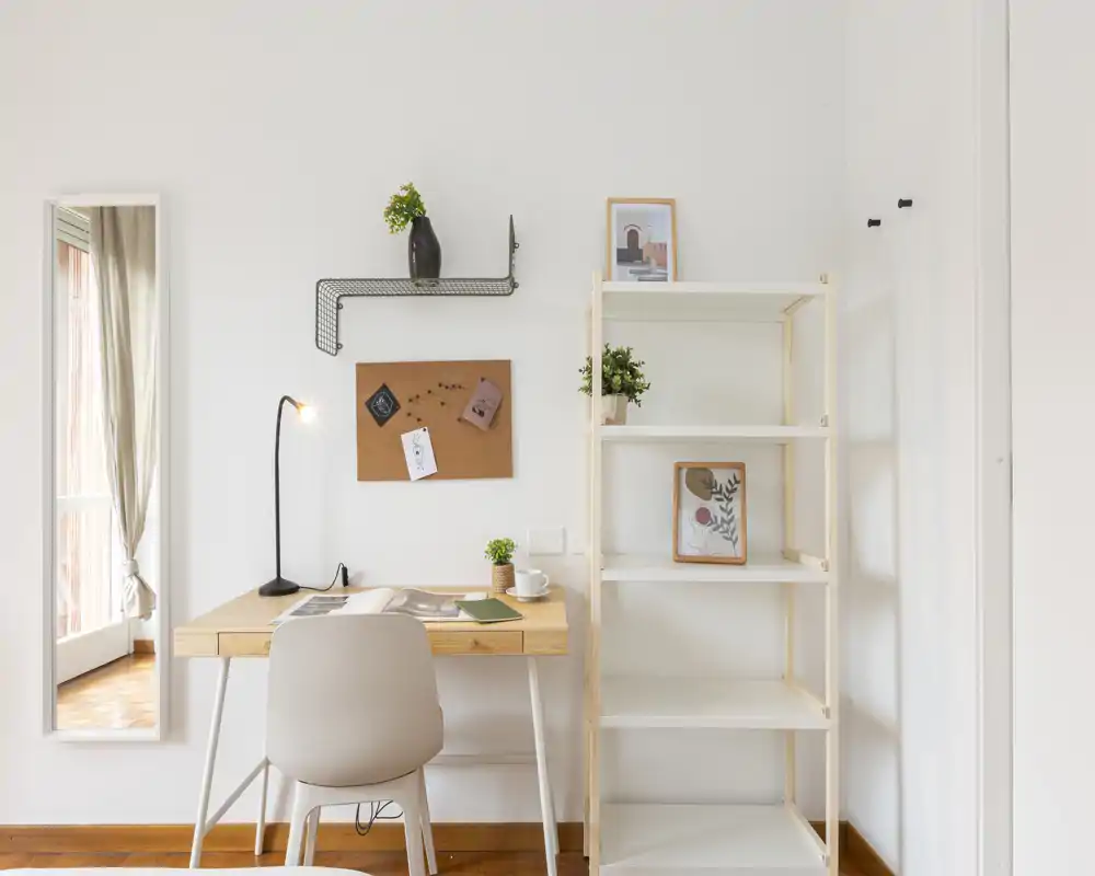 A bright, minimalist living/work corner with a wooden desk, chair, shelving unit and decorative plants—ideal for a compact home office within the living area.