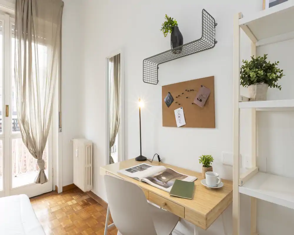 Small bedroom workspace with a wooden desk, chair and a bright window. The scene shows a neatly styled desk with a lamp, open book, cup and plants, and part of the bed visible at the image edge.